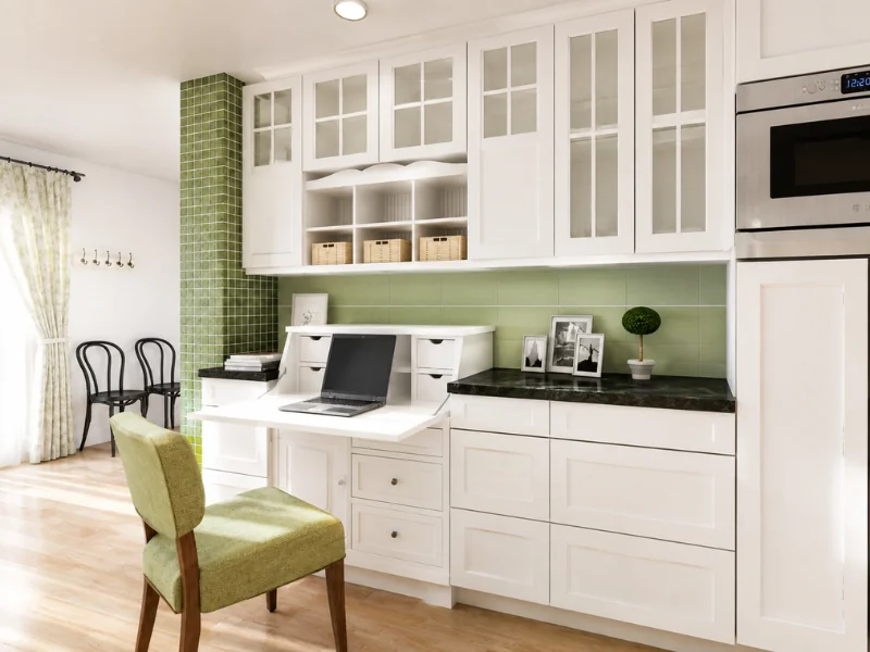 White kitchen cabinetry with a built-in home office, green tile backsplash, glass-front cabinets, laptop, storage baskets, and green chair.