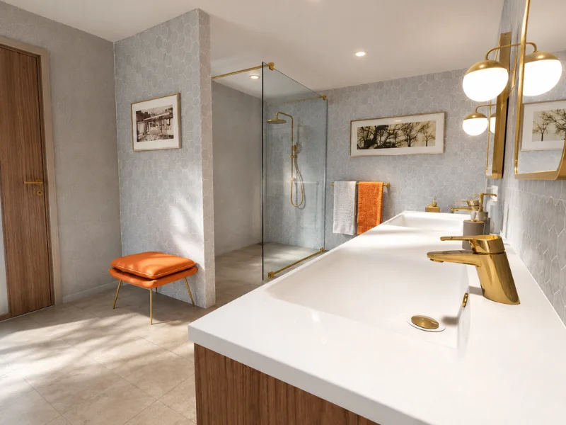 Bright modern bathroom with a long white double vanity, wood cabinetry, brass faucets, glass walk-in shower, orange stool, and pale hexagon tiles.