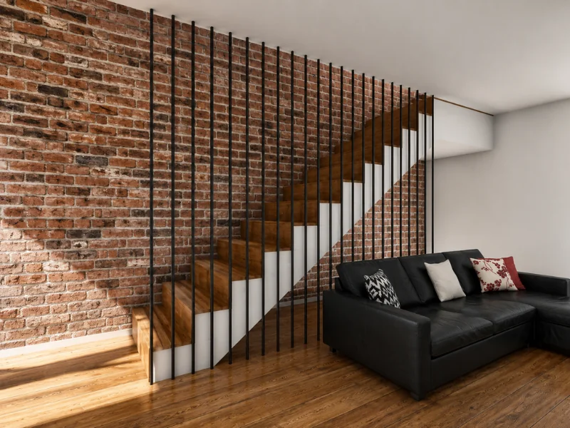 Wood staircase with slim black metal balusters, exposed brick walls, and a black sofa beside the stairs.