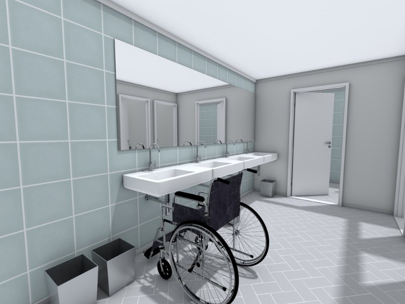 Accessible public restroom with wheelchair positioned at a row of sinks, showing clear knee space, long mirror above, and light blue tiled walls.