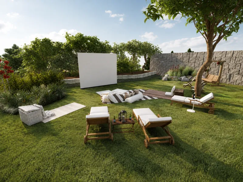 Backyard lawn set up as an outdoor movie area with a projection screen, lounge chairs, floor cushions, and surrounding greenery.