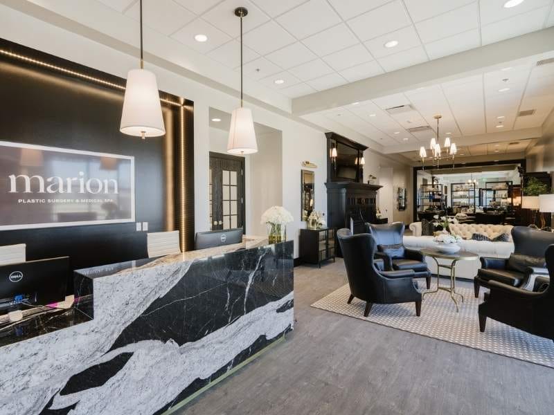 Elegant reception area and waiting room featuring a black marble front desk, modern pendant lights, and comfortable lounge seating. The space combines classic and contemporary design elements with neutral tones and warm lighting.