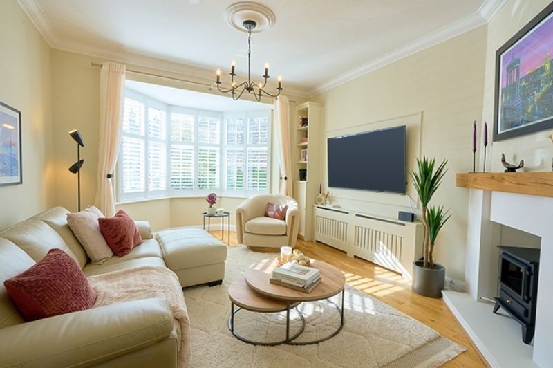 Bright living room with a large bay window, cream sectional sofa, and a round armchair accented with red throw pillows. A round wooden coffee table with books and candles sits on a light rug. A wall-mounted TV is framed by built-in shelves and a radiator cover, while a fireplace with a wooden mantel and potted plant completes the space.