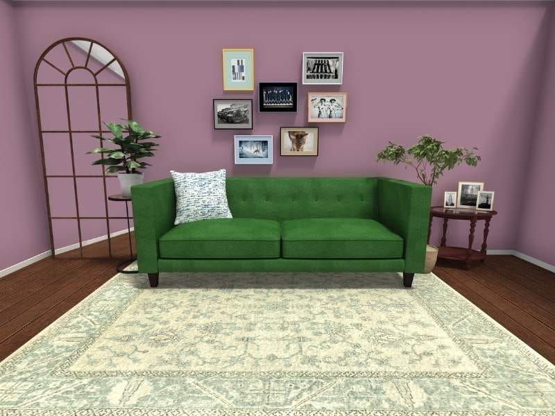 3D Photo of a living room. The camera is pointed directly at a green couch with a blue and white rug underneath it. There is a white and blue pillow on the couch. On each side of the couch there are plants, on one side is a floor mirror and on the other, a side table with three pictures placed on top. Above the couch is a picture wall with seven pictures.