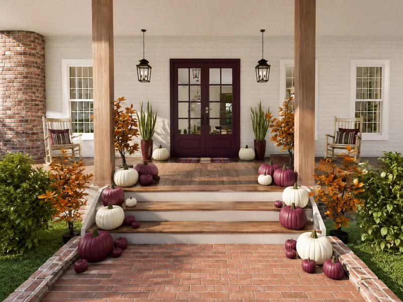 Front porch decorated for autumn with burgundy and white pumpkins, orange-leaved plants, wooden rocking chairs, hanging lanterns, and a dark double front door.