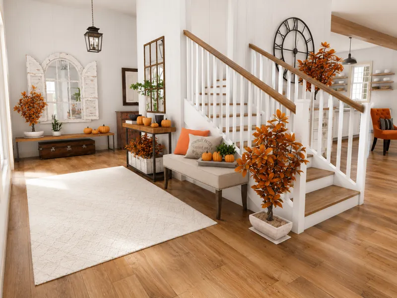Bright entryway decorated for autumn with orange-leaved plants, small pumpkins, a cushioned bench, a large wall clock, and a white staircase with wooden handrails.