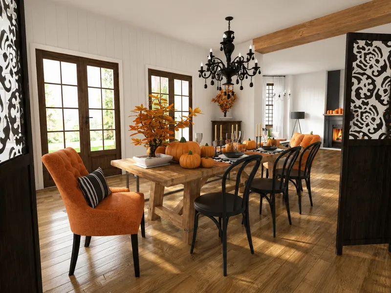 Autumn-themed dining room with a rustic wooden table, orange upholstered chairs, black chairs, pumpkins, orange foliage, a black chandelier, and decorative patterned screens.