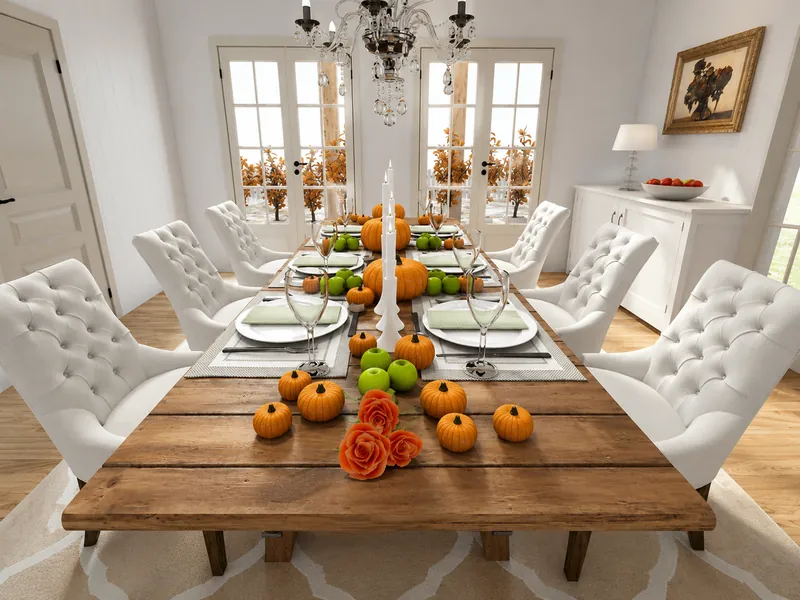Elegant dining room with a rustic wooden table set for eight, white tufted chairs, pumpkins, green apples, candles, orange roses, and a crystal chandelier.