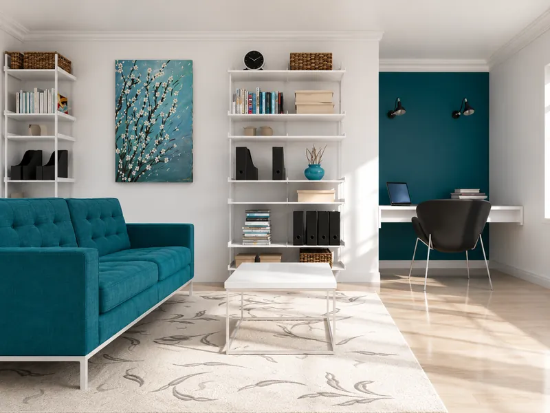 Bright home office and sitting area with a teal sofa, white shelving, floral wall art, a compact desk nook, and a patterned rug.