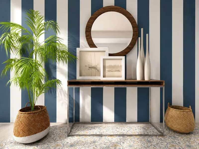 Entryway vignette with navy-and-white striped wallpaper, a slim console table, round wood-framed mirror, framed art, white decorative vases, a potted palm, and woven baskets.