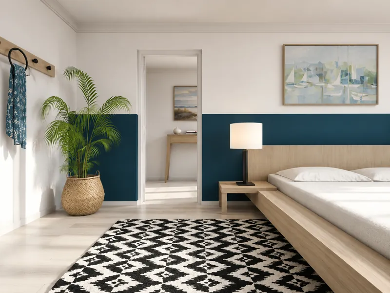 Minimalist bedroom with a light wood platform bed, teal half-wall accent, black-and-white patterned rug, potted palm, bedside lamp, and soft coastal artwork.