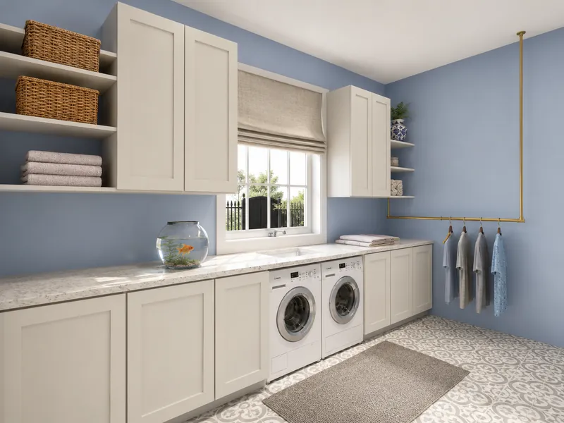 Bright laundry room with soft blue walls, white shaker-style cabinets, open shelving, patterned floor tiles, and a front-loading washer and dryer beneath a window. A brass hanging rail, folded towels, woven baskets, and a small fishbowl complete the space.