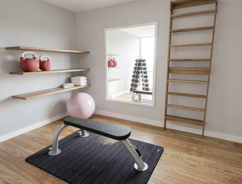 Home gym with a workout bench on a black mat, a pink exercise ball, wooden wall shelves holding kettlebells and towels, a large mirror, and a wooden wall ladder.