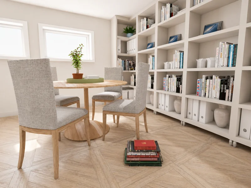Bright library or study room with floor-to-ceiling bookshelves, a round wooden table, four upholstered chairs, potted plants, and books stacked on the wood floor.