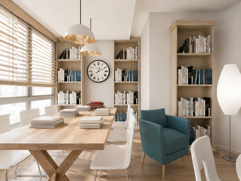 Bright reading and study room with a long wooden table, white chairs, floor-to-ceiling bookshelves, a blue armchair, pendant lights, and a large wall clock.