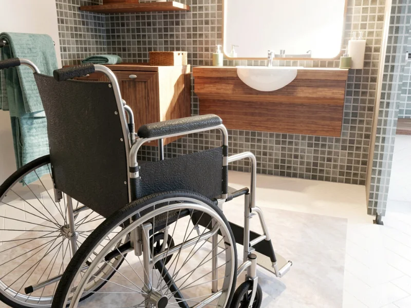 3D rendering of an accessible bathroom with a wheelchair in front of a floating wooden vanity and wall-mounted sink. The room features small gray mosaic wall tiles, open floor space, folded towels, storage cabinets, and a large illuminated mirror.