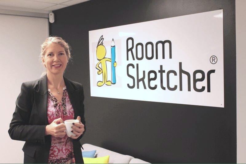 RoomSketcher CSMO Trine standing in an office holding a coffee cup, smiling in front of a wall sign that reads "Room Sketcher" with the brand's yellow mascot holding a blue pencil. The setting is modern and professional, with a dark feature wall and a couch with colorful pillows below the sign.