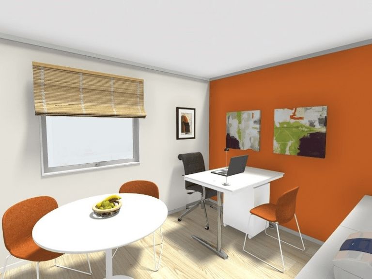 3D rendered image of a modern home office or small meeting room with an orange accent wall. The room features a white desk with a laptop, a black office chair, and an orange visitor chair. A round table with three orange chairs and a fruit bowl is placed near the window, which has a bamboo-style blind. Abstract artwork decorates the walls, and the flooring is light wood.