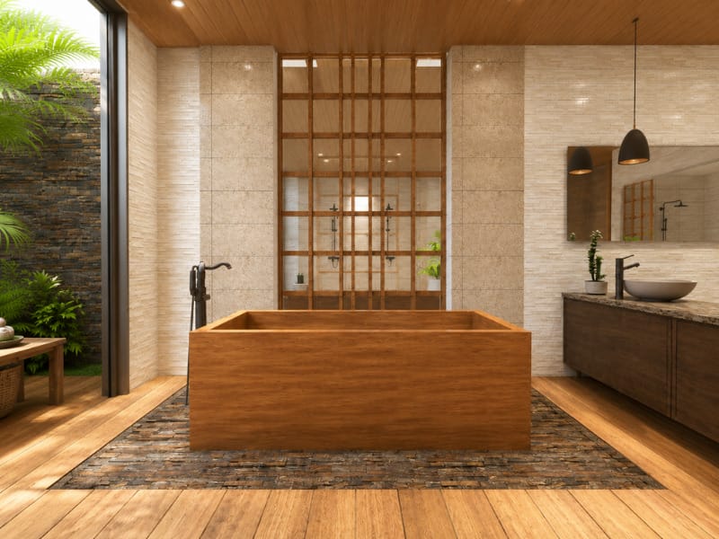 Luxurious bathroom with a rectangular wooden bathtub, natural stone walls, wood-paneled ceiling, double vanity, pendant lights, and a view of lush tropical plants.