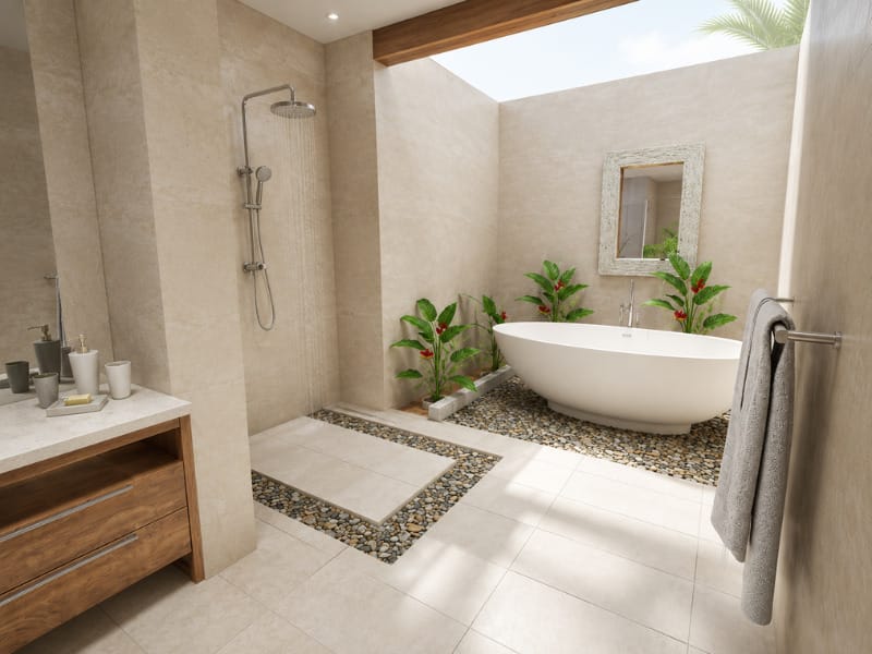 Tropical spa-style bathroom with a freestanding white bathtub, open-air rainfall shower, beige stone tiles, pebble flooring, warm wood vanity, and lush flowering plants.