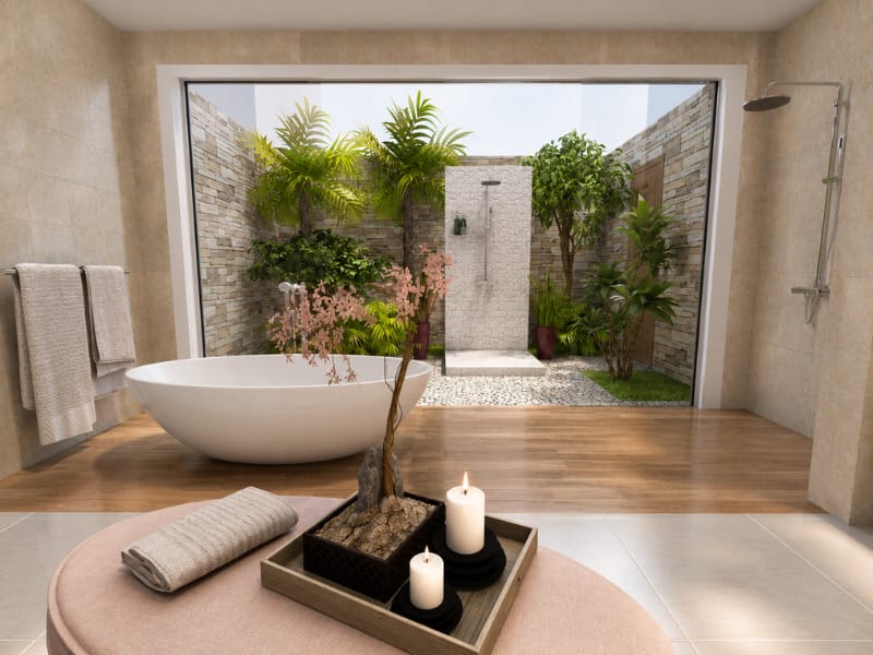 Tropical spa-style bathroom with a freestanding white bathtub, open-air garden shower, natural stone walls, wood-look flooring, candles, towels, and lush greenery.