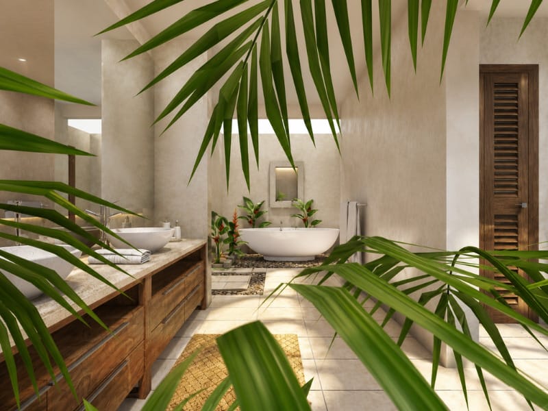 Tropical organic bathroom with a wooden double vanity, vessel sinks, freestanding bathtub, textured neutral walls, natural stone details, louvered wood door, and abundant palm foliage.