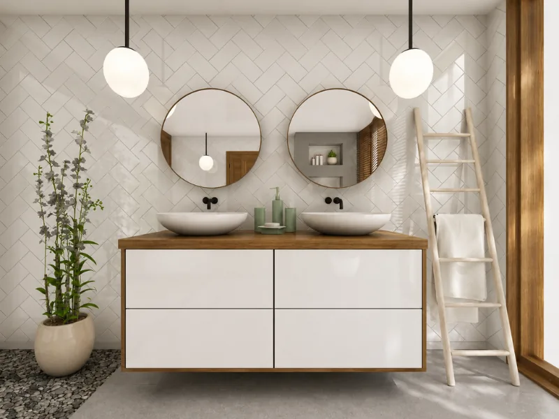 Bright modern bathroom with a floating double vanity, two vessel sinks, round mirrors, globe pendant lights, white herringbone tiles, a towel ladder, and potted greenery.