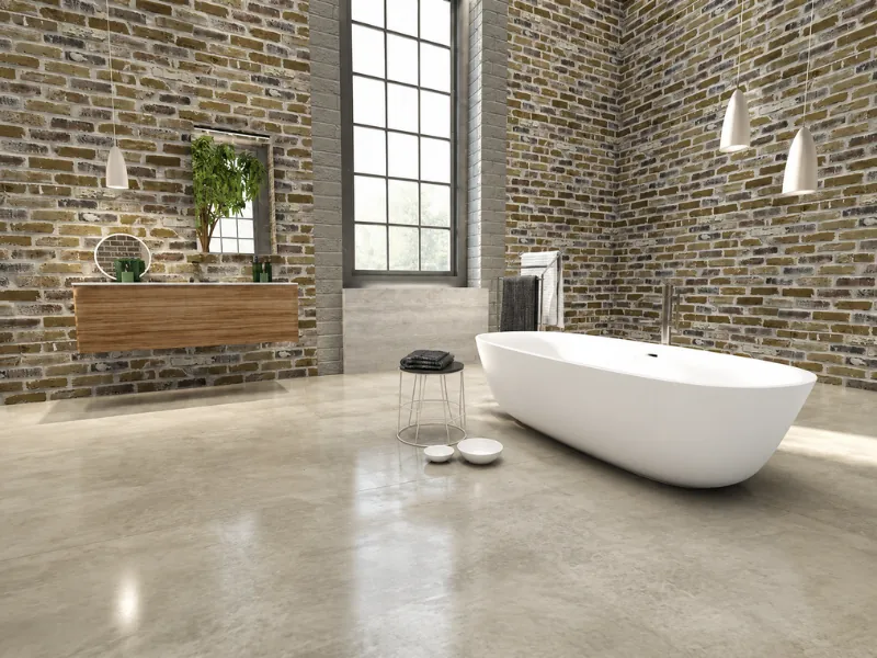 Spacious loft bathroom with exposed brick walls, a white freestanding tub, floating wood vanity, polished concrete flooring, pendant lights, and a tall grid window.