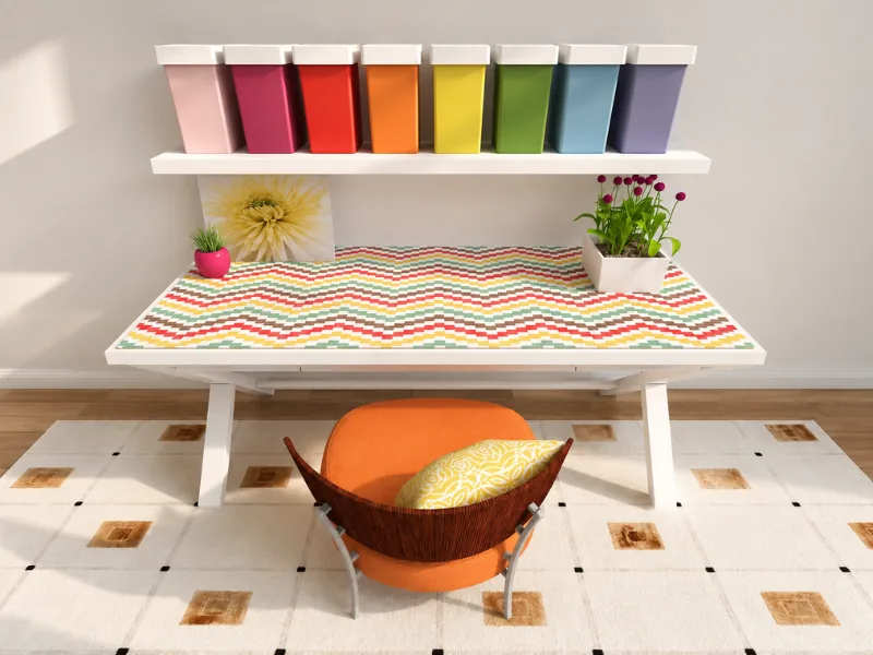 Cheerful home office with a colorful patterned desk, orange chair, rainbow storage bins on a wall shelf, potted plants, and a geometric area rug.