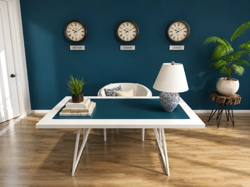 Stylish home office with a deep blue accent wall, three world clocks, a white desk with blue inset, upholstered chair, ceramic lamp, wood flooring, and a potted palm.