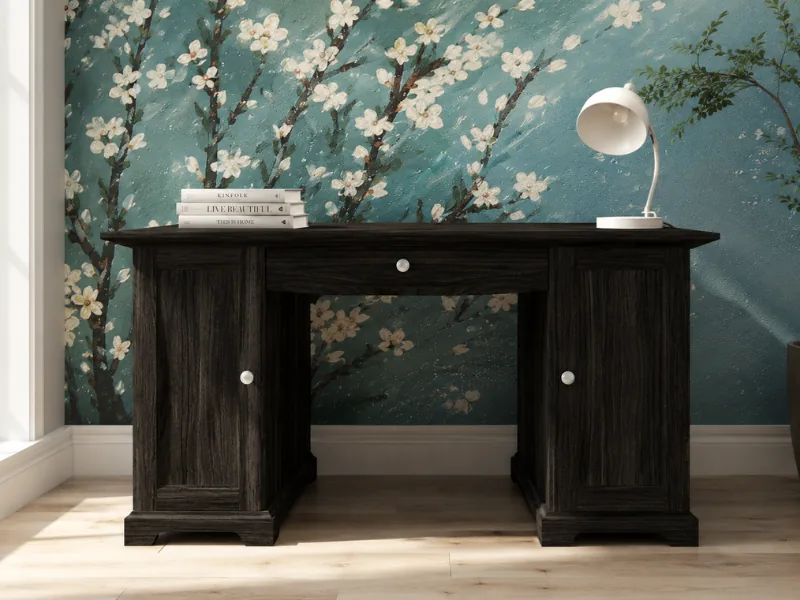 Home office with a substantial black wood desk, white task lamp, stacked books, and a teal floral accent wall featuring blossoming branches.