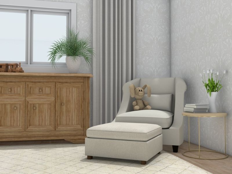Cozy nursery corner with a light gray armchair and matching ottoman, decorated with a stuffed bunny toy. Beside the chair is a small round side table with a vase of white flowers and stacked books.
