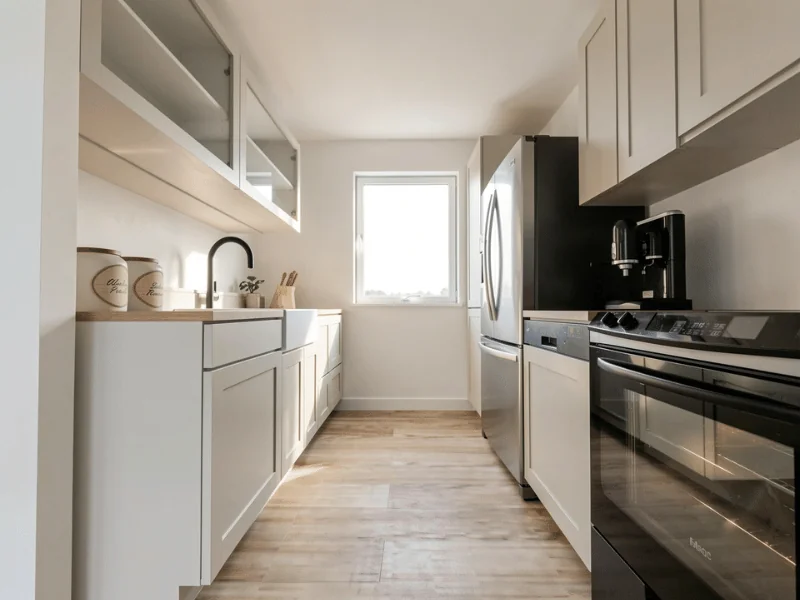 Modern galley kitchen with white shaker cabinets, light wood flooring, stainless steel refrigerator and oven, black faucet, and a bright window at the end.