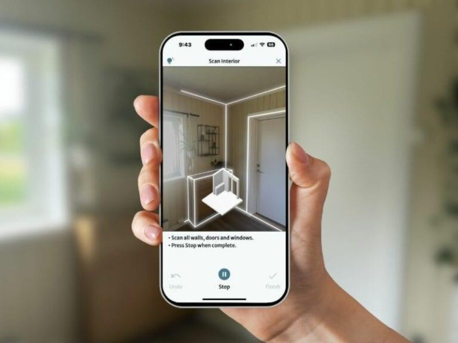 A hand is holding a smartphone running a room scanning application. The app interface shows an augmented reality overlay that outlines the edges of walls, doors, and windows in a room. On the screen, there’s a prompt instructing the user to “Scan all walls, doors and windows” and “Press Stop when complete.” The application appears to be capturing spatial data in real-time, using 3D visualization to generate an interior layout or floor plan. The interface also includes buttons for “Undo,” “Stop,” and “Finish,” indicating the scanning process is interactive and user-controlled.