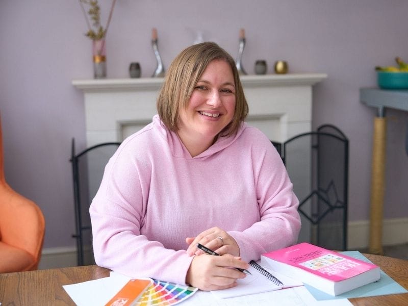 Indretningsarkitekt Debbie Bryant i pink hættetrøje sidder ved et bord med designmaterialer, herunder en farveprøvebog, papirer og en pink bog, i et hyggeligt rum med pejs i baggrunden.