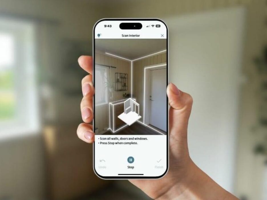 En hånd holder en smartphone, der kører en rumscanningsapp. Appens grænseflade viser et lag med udvidet virkelighed (augmented reality), som markerer kanterne på vægge, døre og vinduer i et rum. På skærmen vises en instruktion, der beder brugeren om at "Scanne alle vægge, døre og vinduer" og "Tryk Stop, når du er færdig." Applikationen ser ud til at indfange rummet i realtid ved hjælp af 3D-visualisering for at generere et interiøroversigt eller en plantegning. Grænsefladen inkluderer også knapperne "Undo", "Stop" og "Finish" på engelsk, hvilket viser, at scanningsprocessen er interaktiv og brugerstyret.
