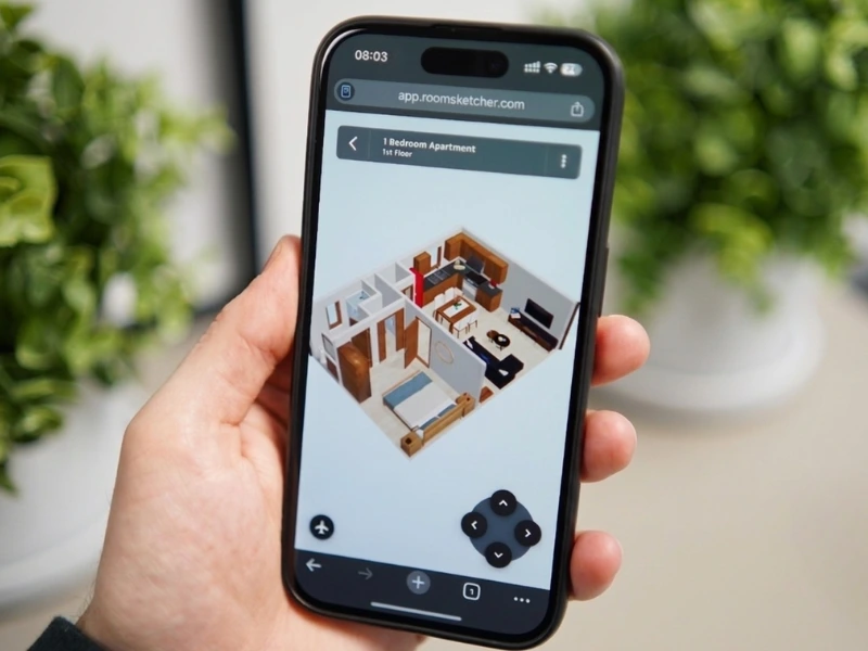 En hånd holder en smartphone, hvor en 3D plantegning vises i RoomSketcher i browseren. Skærmen viser en lejlighed med stue, køkken og soveværelse set i Live 3D, med navigationskontroller nederst.