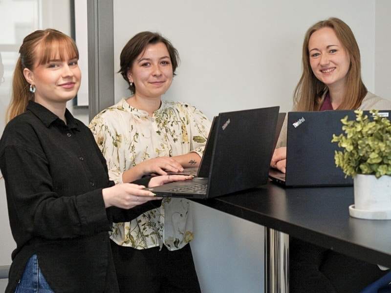 Das Kundenserviceteam von RoomSketcher steht um einen hohen schwarzen Tisch herum, jeder arbeitet an einem Laptop und lächelt in die Kamera.